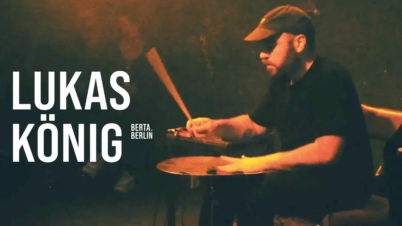 Lukas König assis, la main gauche à plat sur une cymbale, une baguette dans la main droite, son nom en gros sur la gauche.